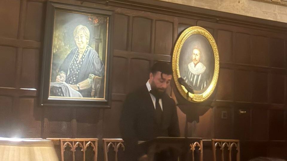 Emre Turan at the lectern in Exeter College hall, Oxford, portraits on the panelled wall behind him